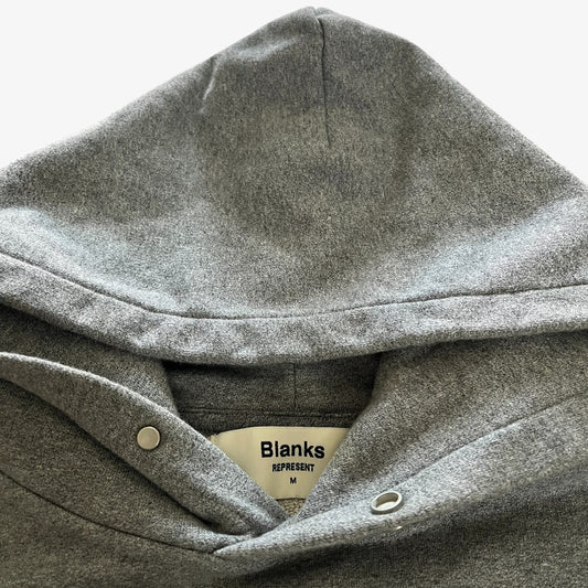 Close-up of a heather grey pullover hoodie hood with snap-button closure and a visible Represent Blanks brand label.