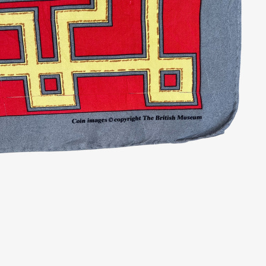 A close-up of a square scarf or fabric corner with a red field, yellow and blue geometric border pattern, and a grey border. Small black text near the bottom reads 'Coin images copyright The British Museum'.