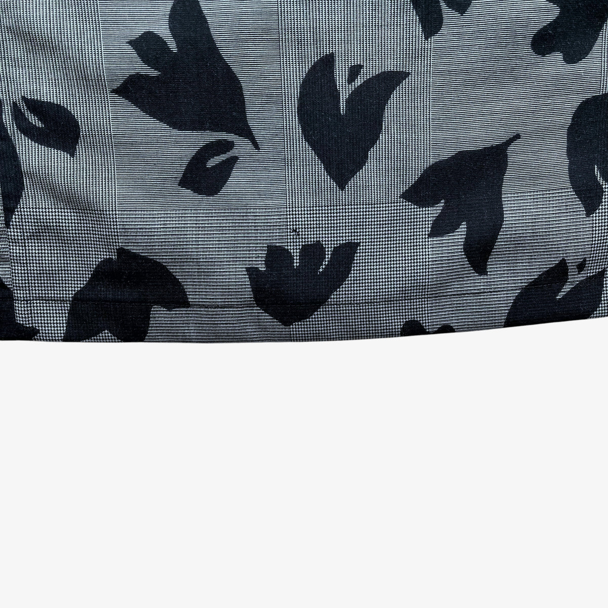 A close-up of a grey and black patterned fabric, showing a fine grid texture with large black abstract leaf on a grey background.