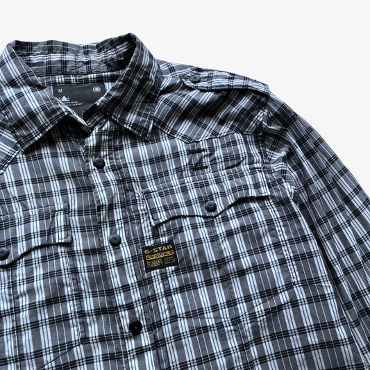 A close-up of a G-Star men's plaid button-down shirt in black, white, and grey, featuring a chest pocket with a flap and a visible G-Star brand patch.