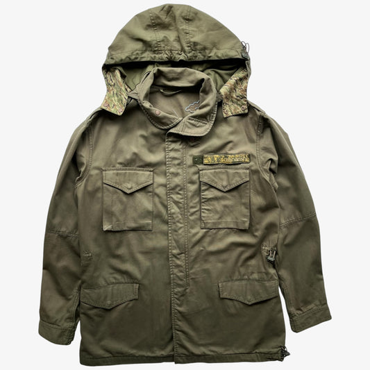 A Khaki green military-style Maharishi hooded jacket with camouflage lining inside the hood, featuring two front flap pockets and a small embroidered patch on the chest, shown against a plain white background.