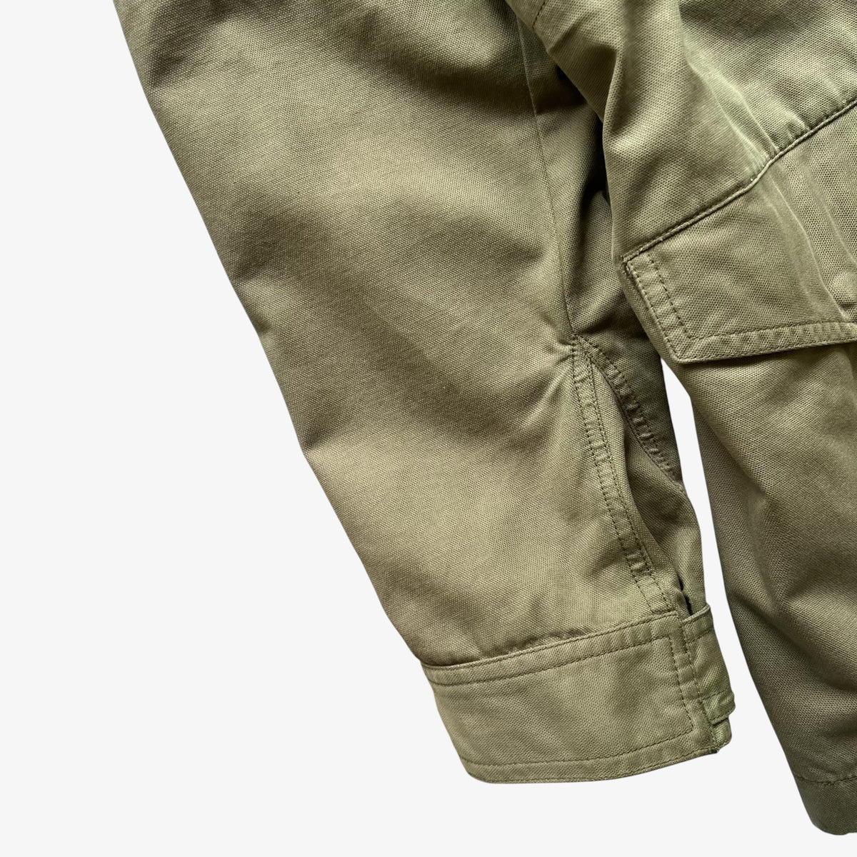 Close-up detail of olive green canvas utility pants showing the sleeve cuff, pocket seam, and fabric texture against a plain white background.