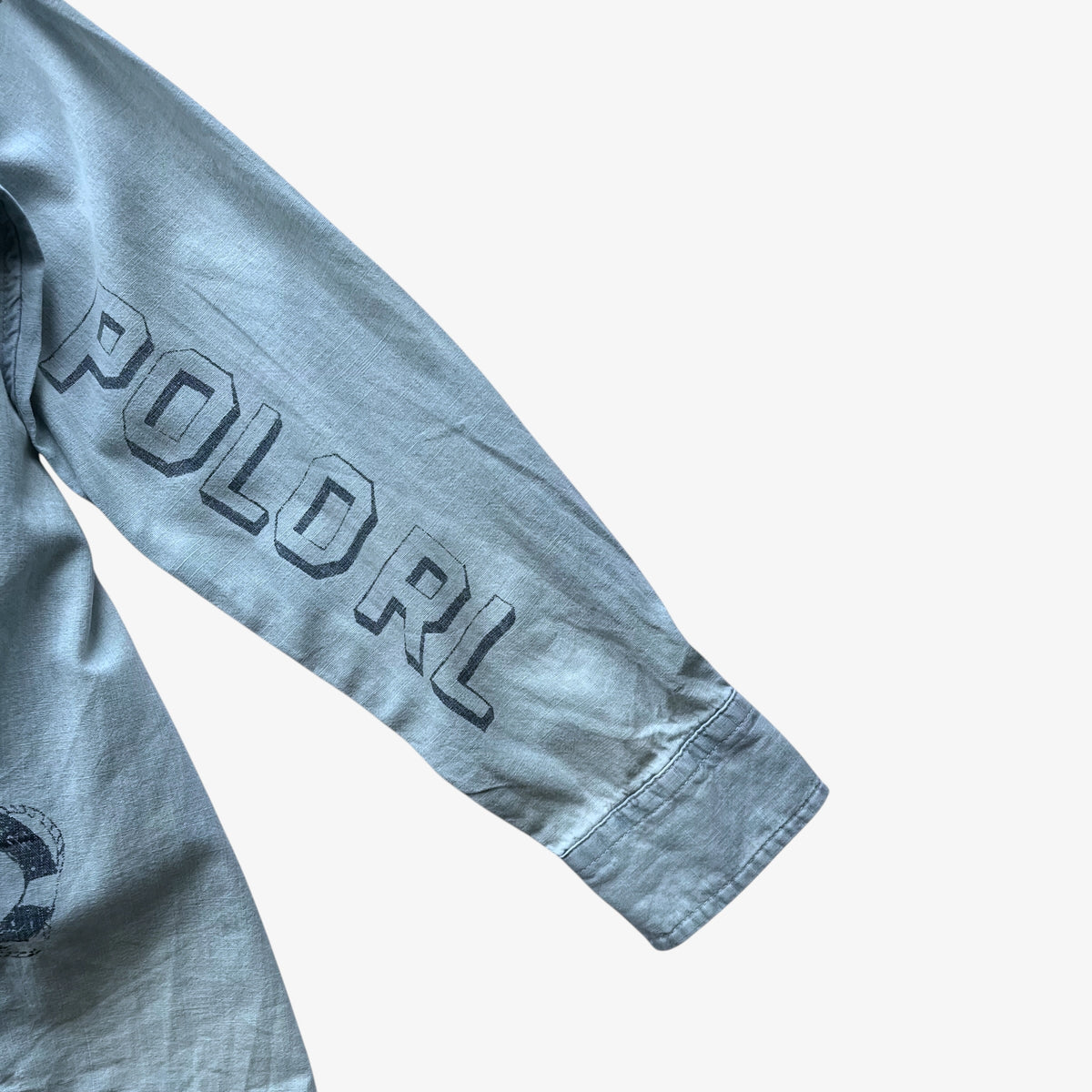 A close-up of a light blue denim long-sleeve shirt showing the left sleeve with 'POLO RL' printed in dark grey block letters and a small circular graphic near the lower left cuff.