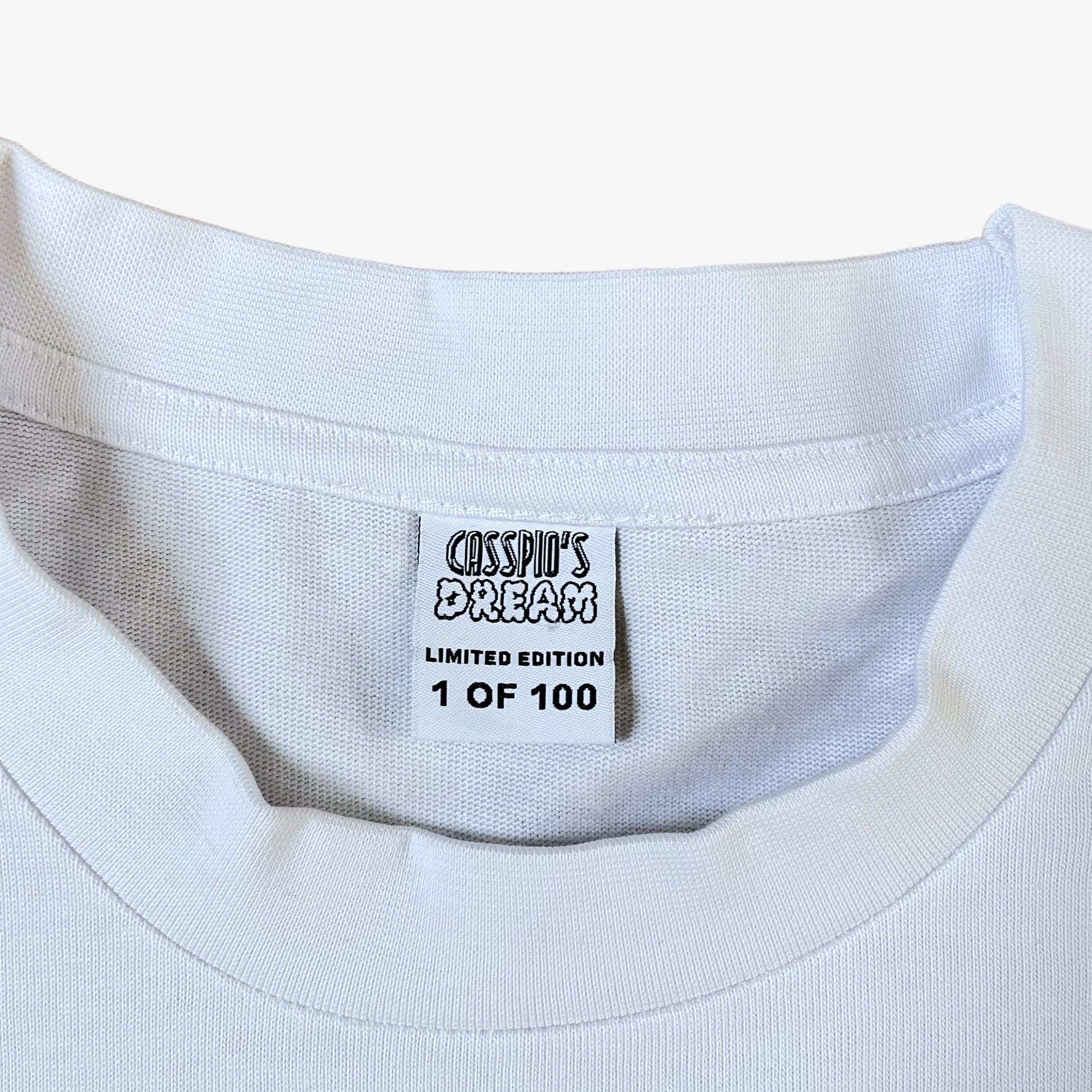 Label of a white 100% organic cotton t-shirt with a graphic print featuring a Banksy styled cartoon character, police dogs and text that reads 'DREAMERS NEVER STOP creating'.