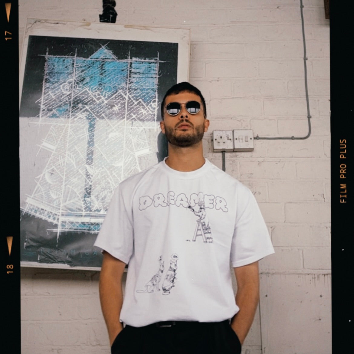 Modelled photo of a white 100% organic cotton t-shirt with a graphic print featuring a Banksy styled cartoon character, police dogs and text that reads 'DREAMERS NEVER STOP creating'.