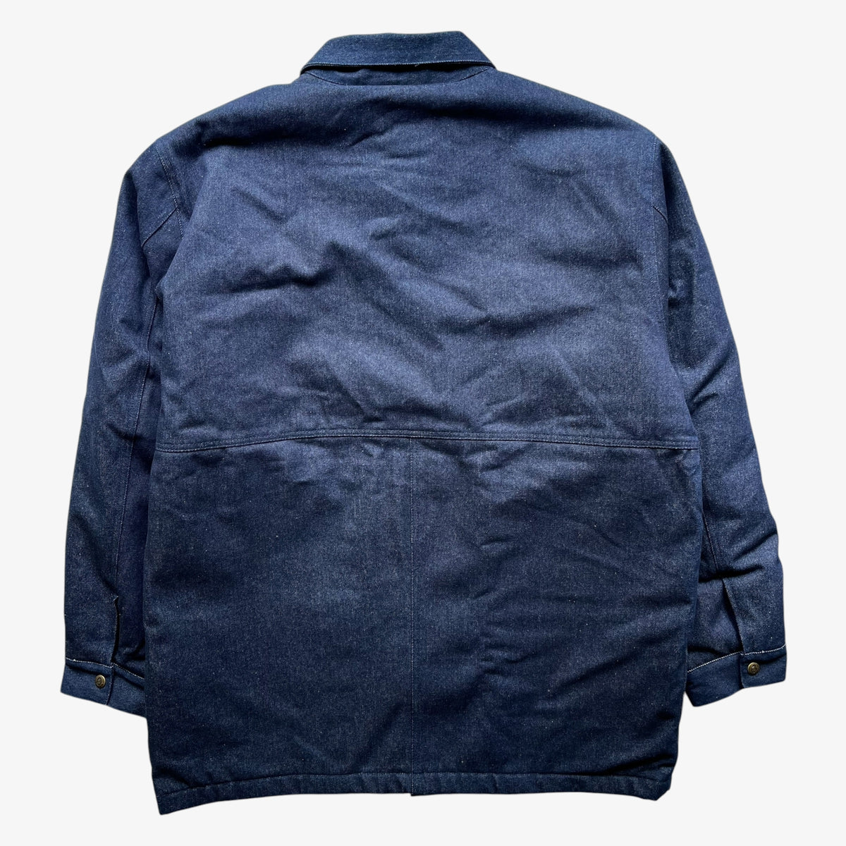 The back view of a blue denim canvas workwear jacket with buttoned cuffs, shown flat against a light off-white background.