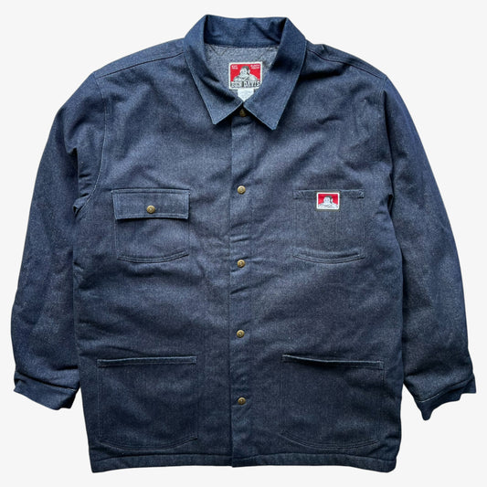 A Ben Davis blue canvas denim workwear jacket with button closures, flap pockets, and a small red and white logo patch on the chest, displayed flat against a light grey background.