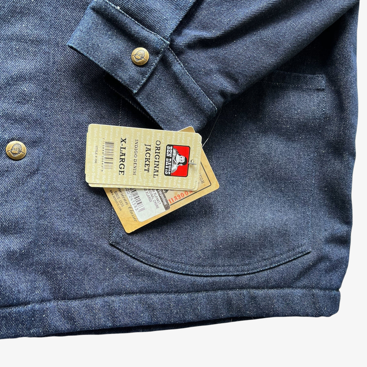 Close-up of a dark blue denim jacket with brass buttons and a paper hangtag reading Ben Davis X-Large.