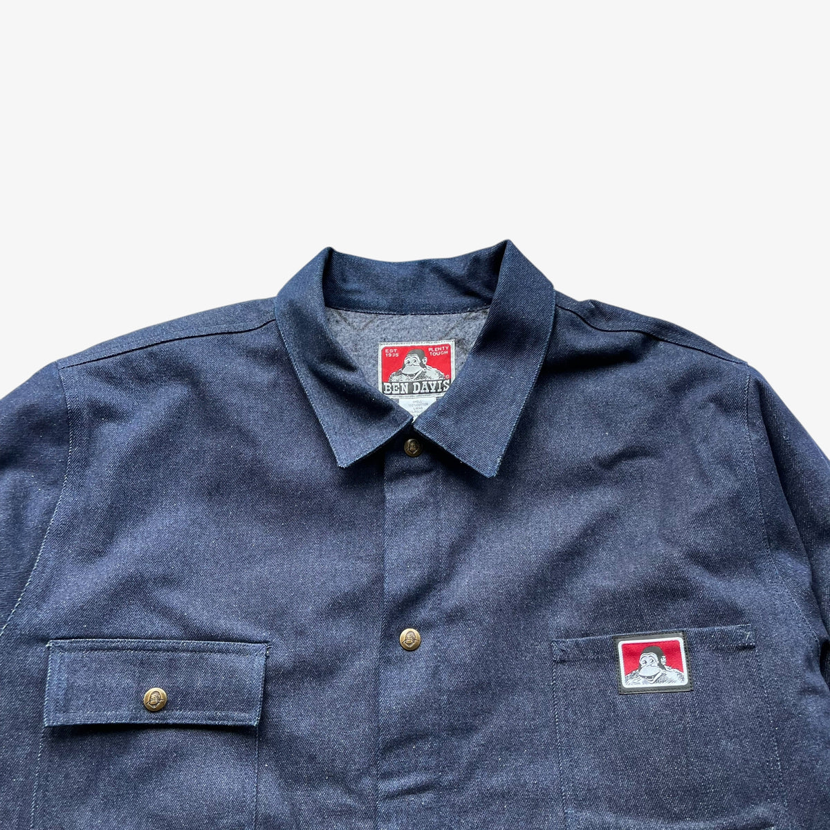 A close-up of a blue denim workwear jacket with two buttoned chest pockets and a small red-and-white brand patch on the chest pocket. The collar label also shows the brand name.