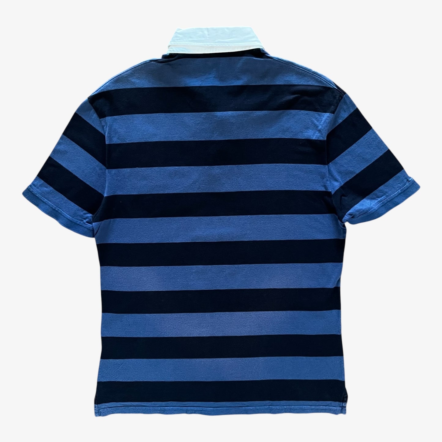 A short-sleeve polo shirt shown from the back, featuring horizontal stripes in shades of blue and navy, with a white collar, on a plain white background.
