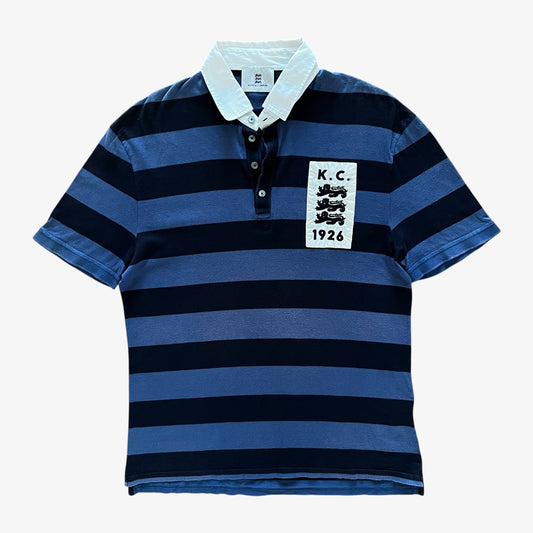 A short-sleeve polo shirt with horizontal navy and medium blue stripes, featuring a light blue collar and a white rectangular patch on the left chest with 'K.C.