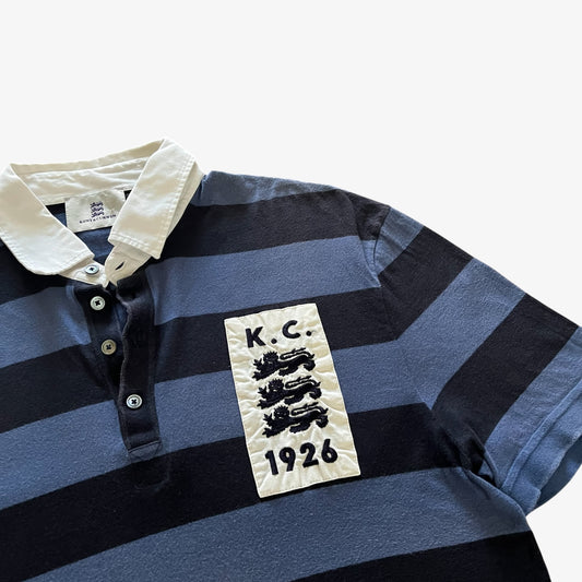 A close-up of a striped short-sleeve polo shirt with navy, blue, and gray horizontal bands, a cream collar, and a cream rectangular chest patch with black text reading 'K.C.