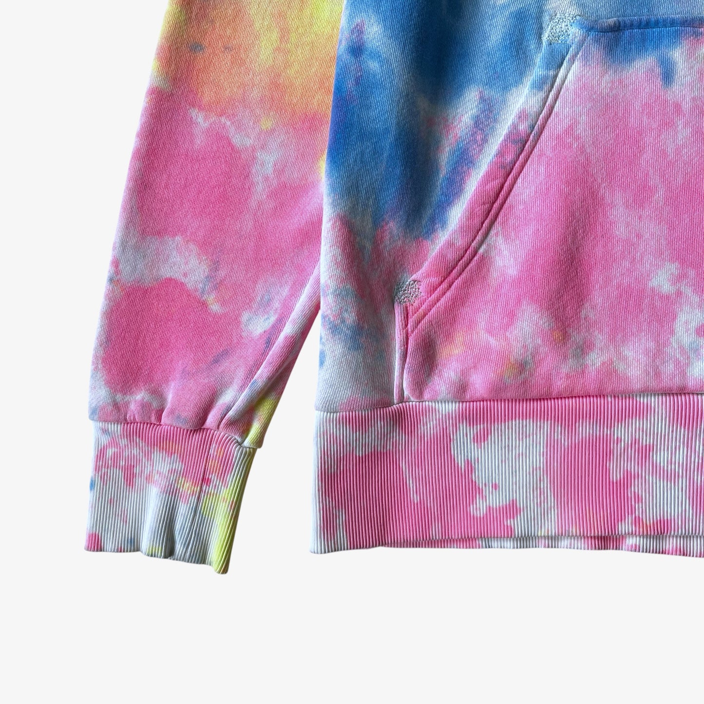 Close up of a tie dye hoodie with pink, blue, and yellow patterns on a white background.