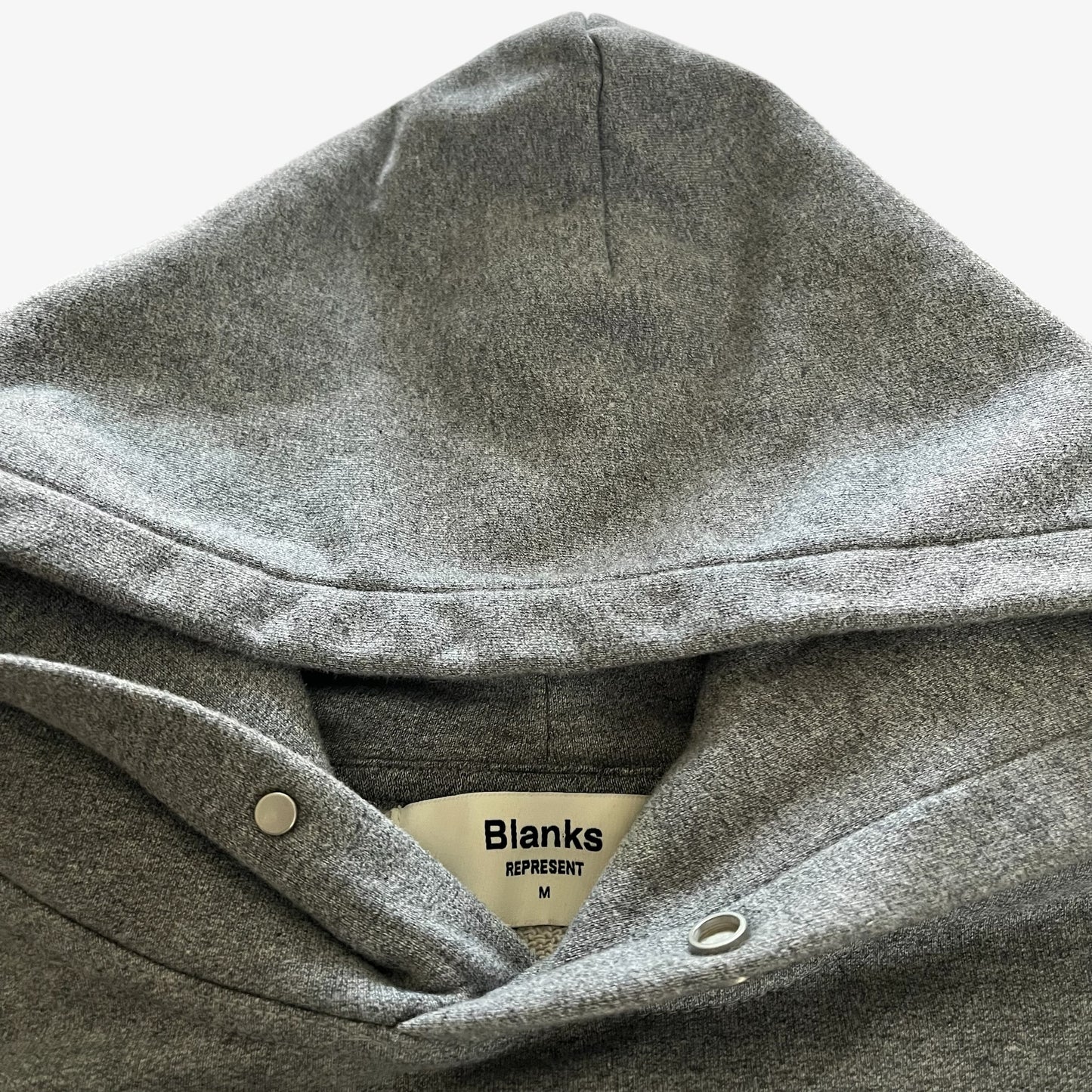 Close-up of a heather grey pullover hoodie hood with snap-button closure and a visible Represent Blanks brand label.