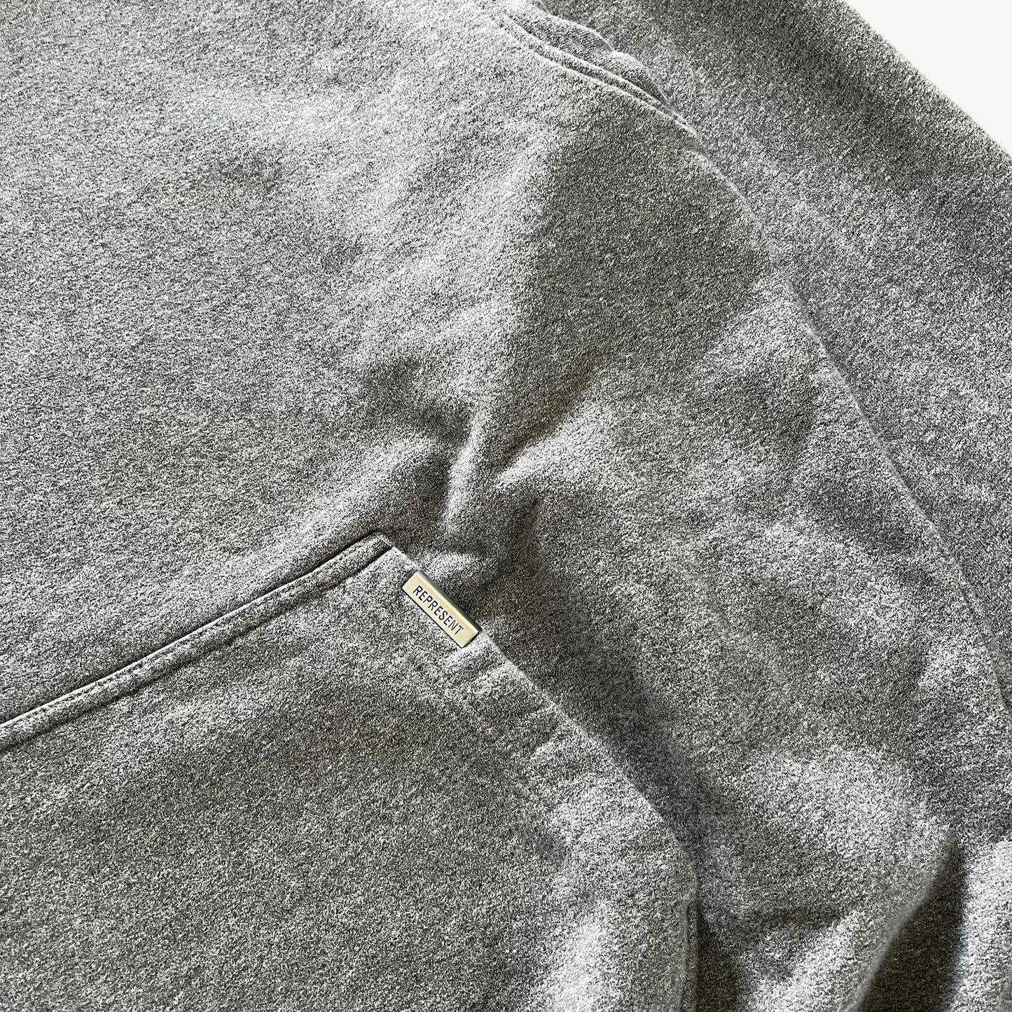 Close-up of a heather grey sweatshirt showing the fabric texture, a pocket seam, and a small silver metal brand tag that reads Represent.