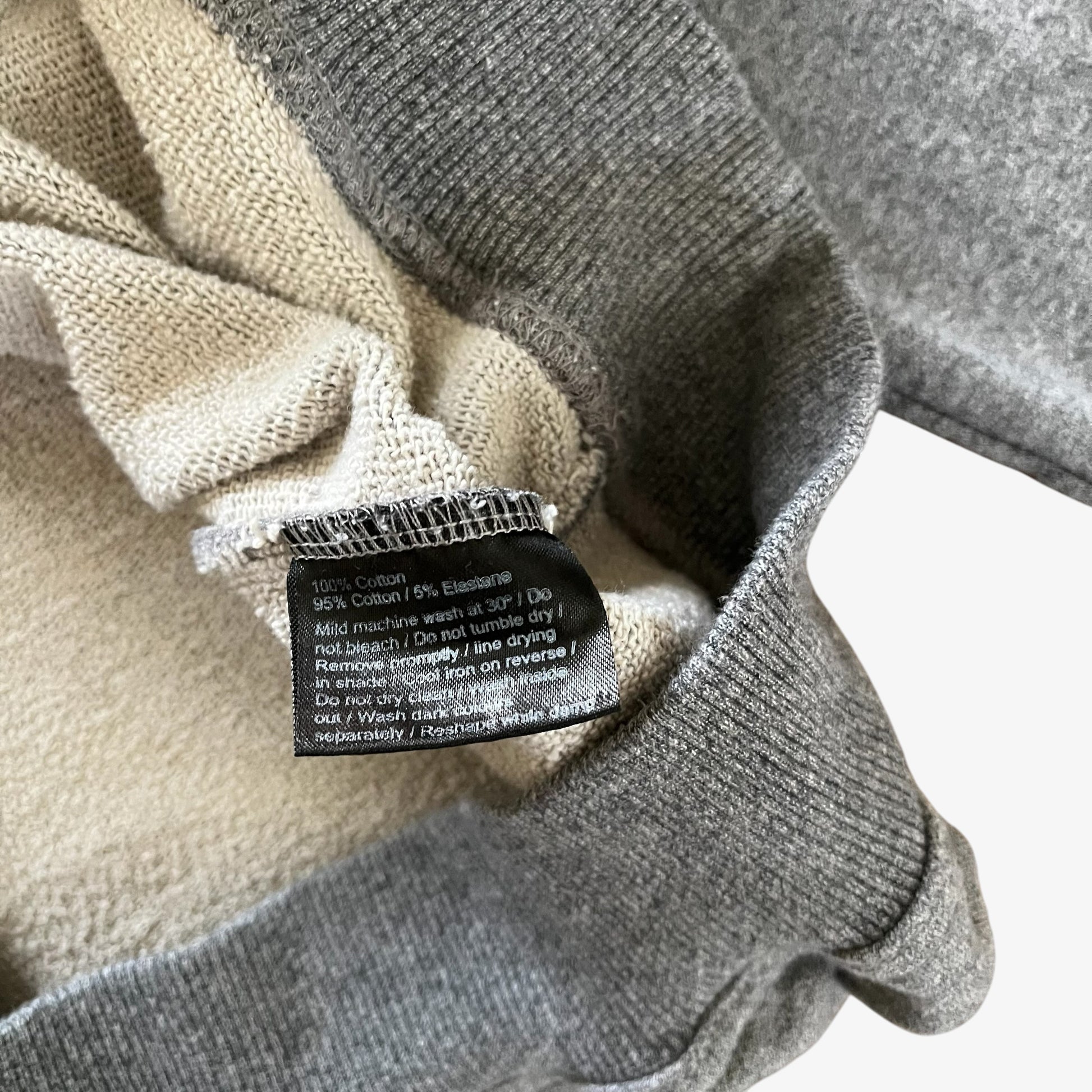 Close-up of a grey ribbed knit hoodie with a cream inner lining and a black sewn-in care label showing cotton composition.