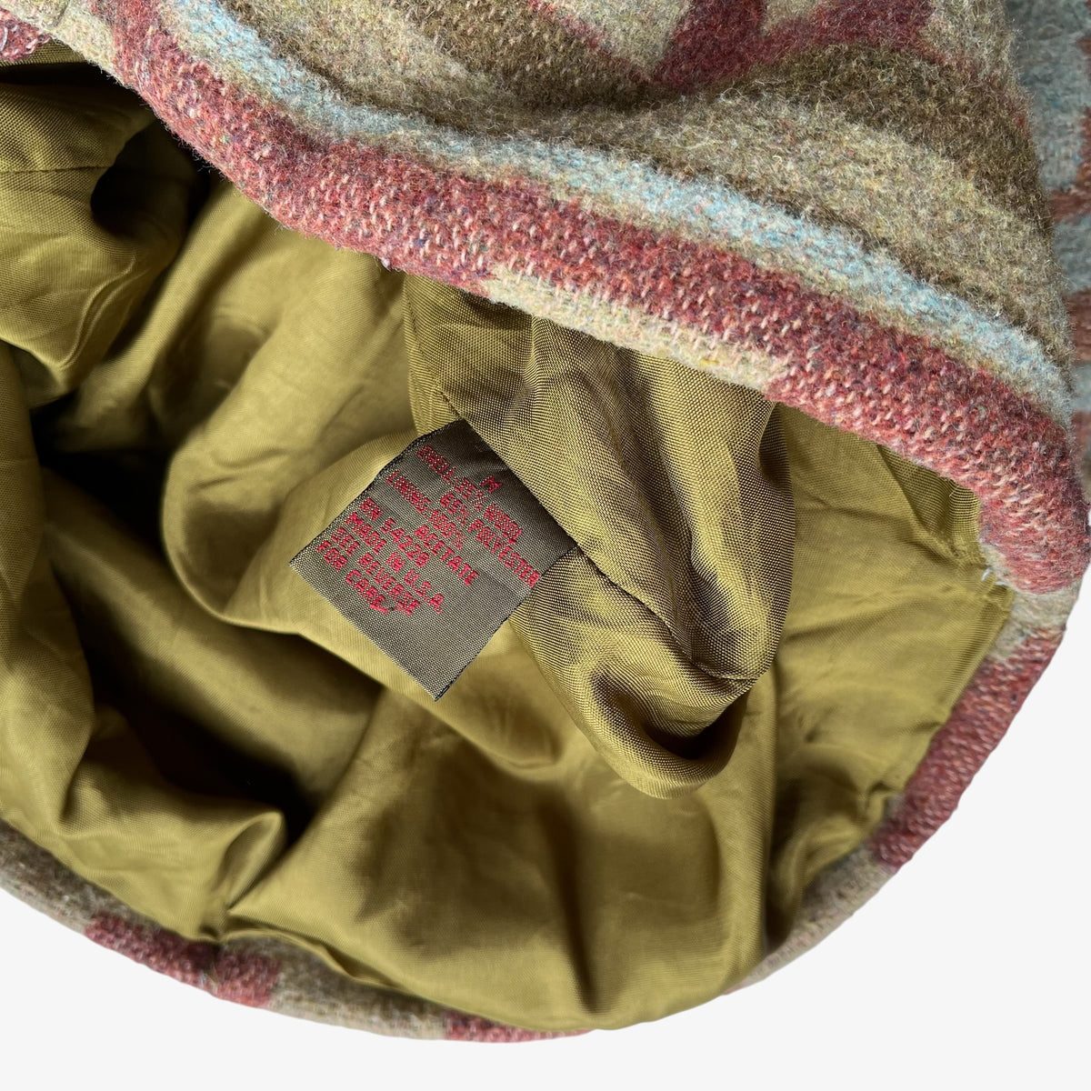 Close-up of a wool jacket with a striped pattern in beige, brown, dusty pink, and light blue, featuring an olive green satin inner lining and a sewn-in brand label.