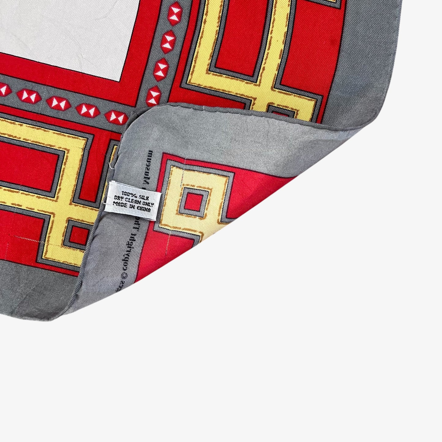 A close-up of hand rolled edges of a square scarf with a geometric pattern in red, yellow, grey, and black, with one corner folded over to show the silk material label.