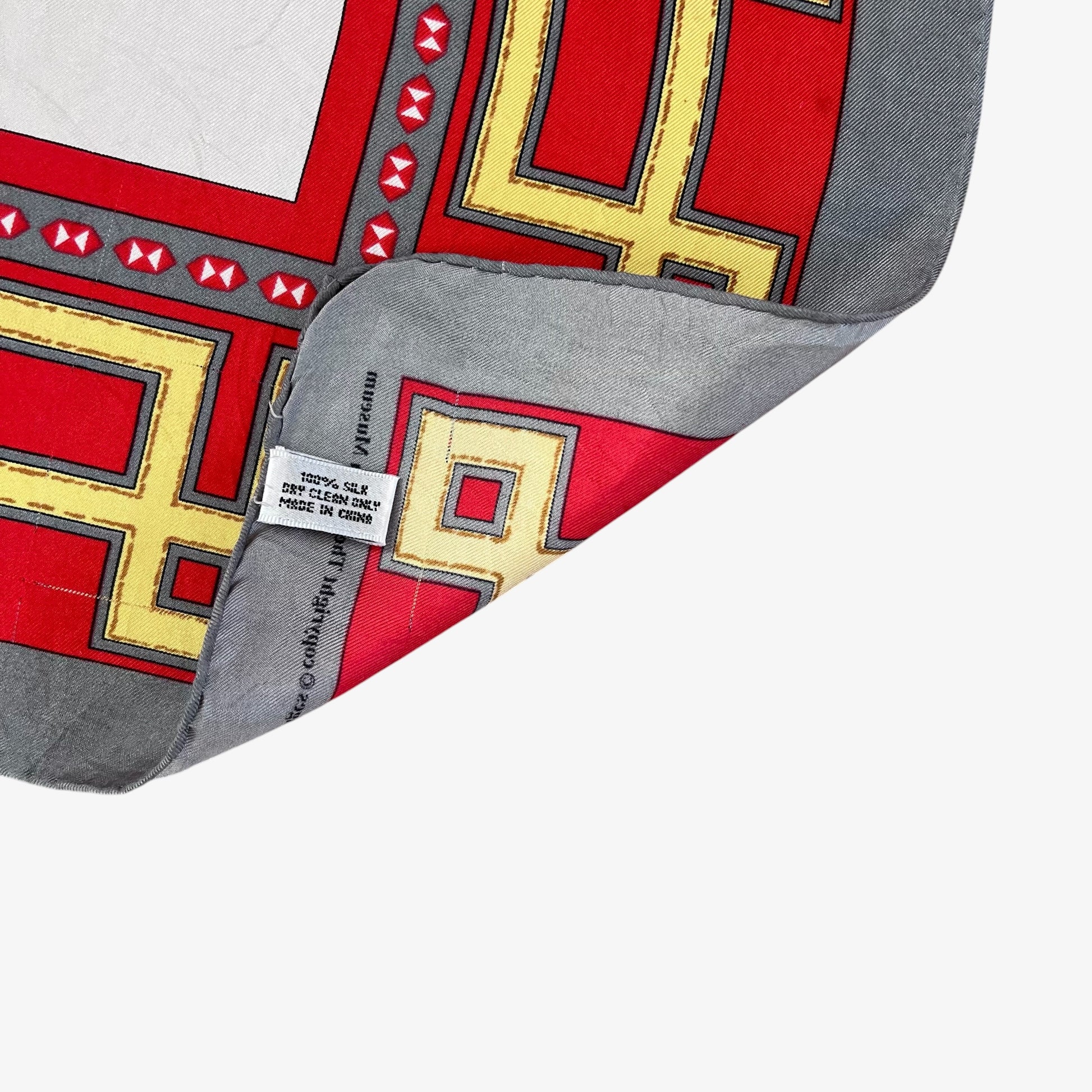 A close-up of hand rolled edges of a square scarf with a geometric pattern in red, yellow, grey, and black, with one corner folded over to show the silk material label.