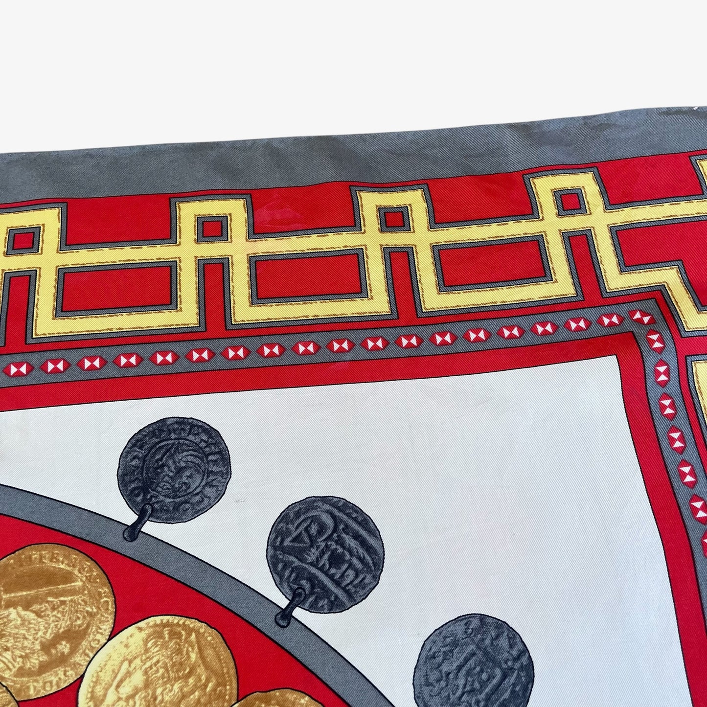 A close-up of a patterned scarf showing a red border with a gold coin key motif, grey trim, and decorative medallion details on a white background.