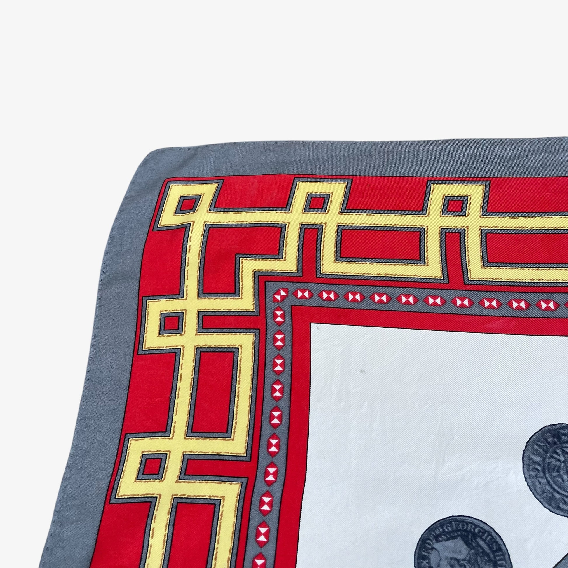 A close-up of a square scarf showing its corner and border details. The design features a grey base with a red border decorated with a gold geometric pattern and small red bow accents, with a white centre containing partial black coin illustrations.