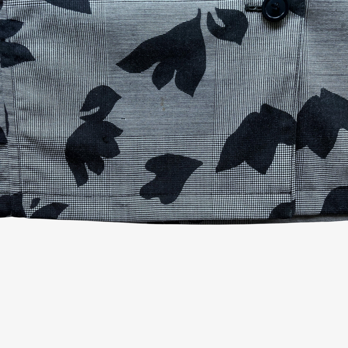 Close-up of a grey and black patterned fabric garment, showing a fine textured weave, a button closure, and a large abstract black leaf or floral motif on a grey background.