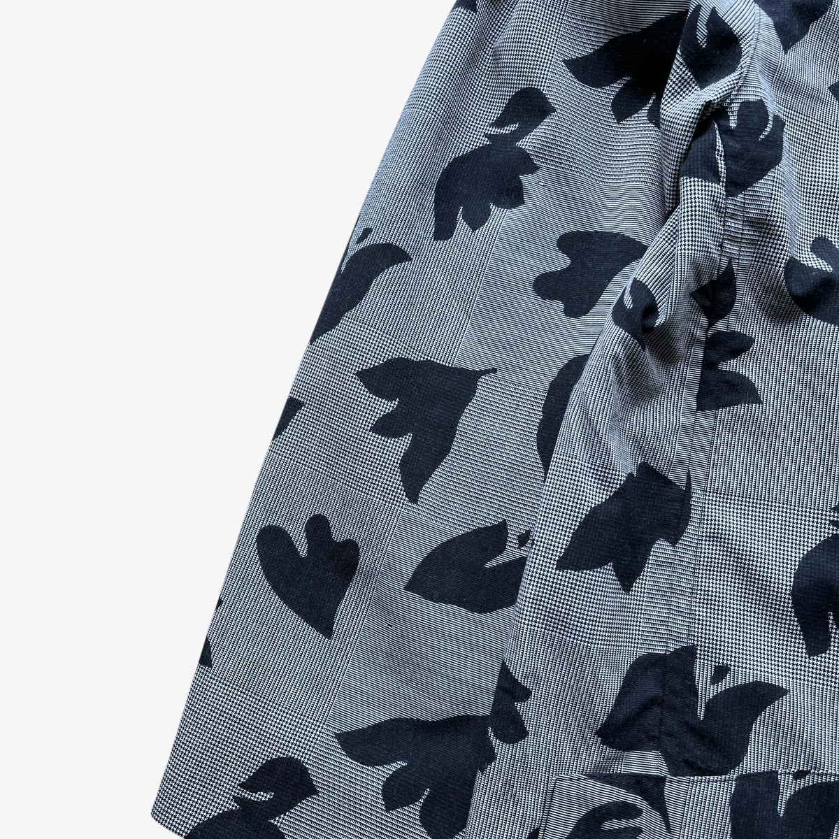 Close-up of a lightweight woven fabric with a fine grey and white micro-check pattern overlaid with large dark navy abstract leaf-like silhouettes, shown on a plain white background.