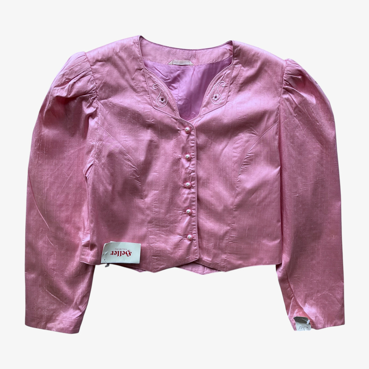 A vintage women's pink trachten jacket with long puffed sleeves, a split neckline, and a button front, laid flat against a light gray background. A small brand tag reading 'Heller Salzburg' is visible near the lower left front.
