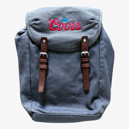 A grey canvas backpack with brown leather straps and silver buckles, featuring a red embroidered Coors beer logo on the front flap.