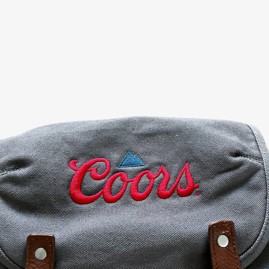 Close-up of a grey fabric bag with red embroidered Coors beer script and a small blue triangle logo, plus brown leather strap hardware visible at the bottom.