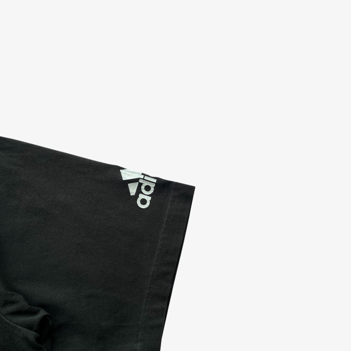 Close-up of a vintage Adidas black t-shirt sleeve corner with a white Adidas logo and trefoil mark on the sleeve, set against a plain white background.