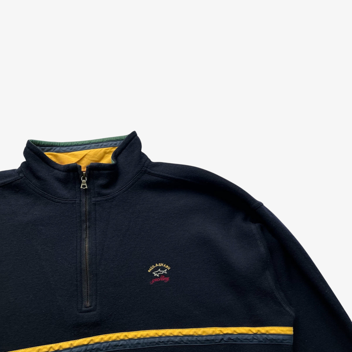 Close-up of a navy wool quarter-zip fleece with a yellow inner collar, yellow horizontal stripe across the chest, and a small embroidered Paul and Shark logo on the left chest, shown on a light grey background.