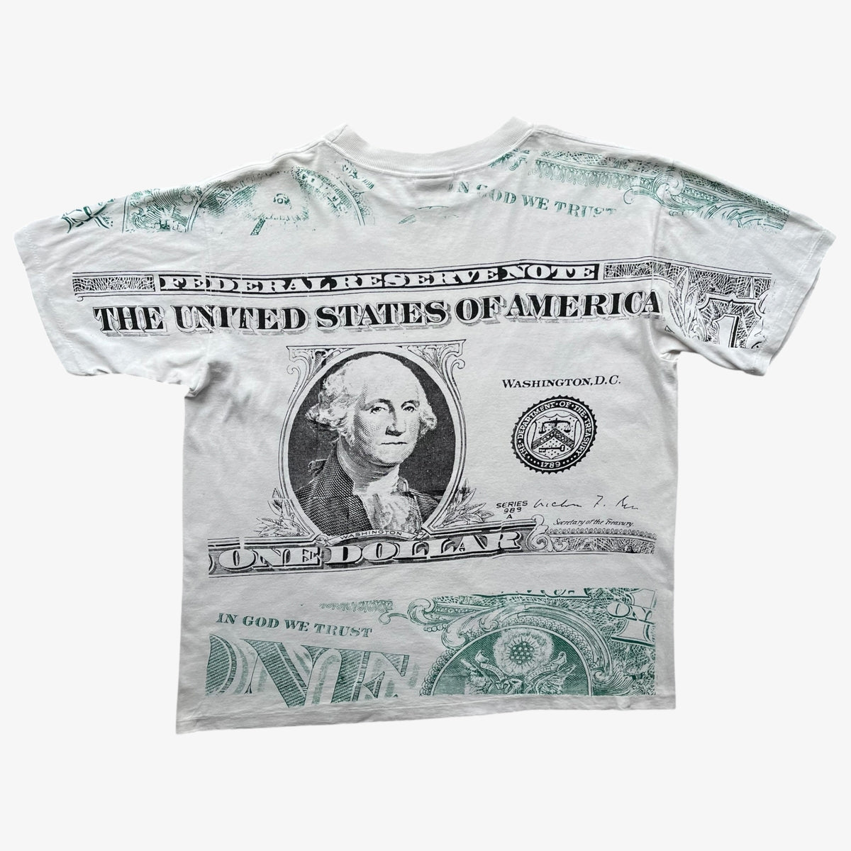 A white graphic t-shirt featuring a detailed print of a US one-dollar bill, including George Washington's portrait, 'THE UNITED STATES OF AMERICA' text, and green security-line details on a light grey background.