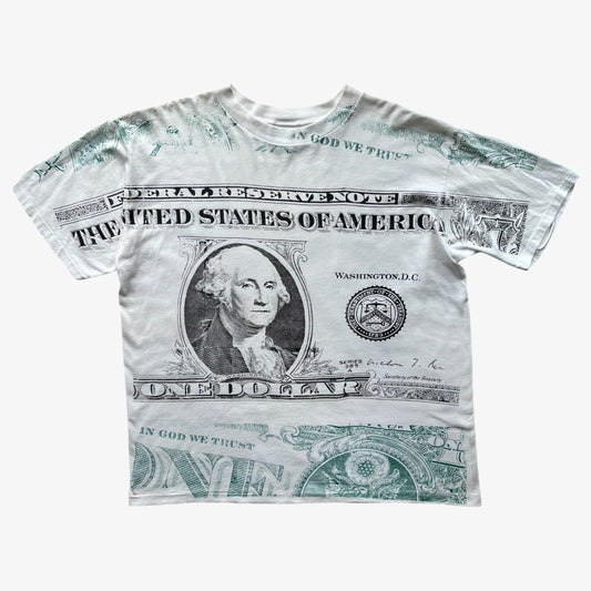 A vintage 90s white short-sleeve t-shirt featuring a detailed graphic print of a US one hundred dollar bill, including George Washington's portrait, 'THE UNITED STATES OF AMERICA' text, and 'IN GOD WE TRUST' printed across the sleeves.