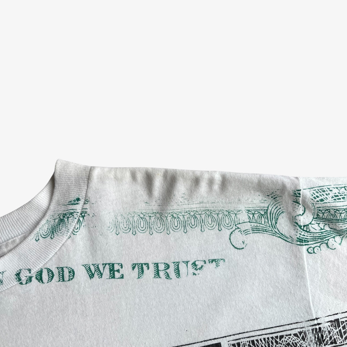 Close-up of a vintage 90s white t-shirt featuring a green printed design inspired by US dollar bill patterns, with the text 'GOD WE TRUST' visible on the front.