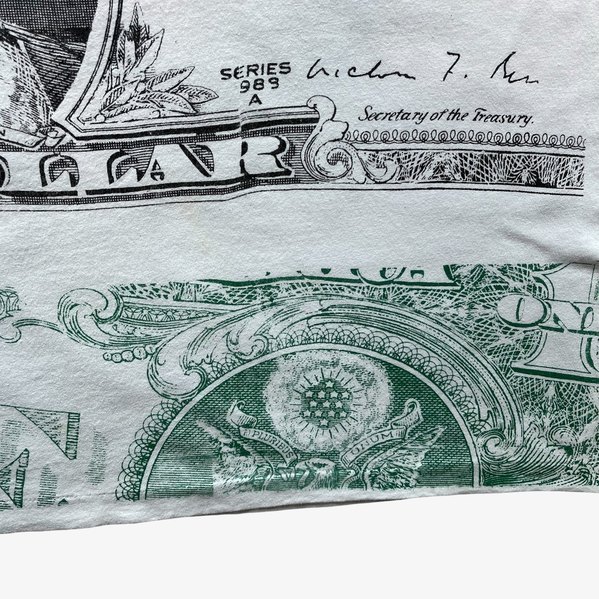 A close-up of a vintage 90s white t-shirt, printed with a detailed graphic of a US one-dollar bill, featuring green ink ornamental details and black text including 'SERIES 1989' and 'Secretary of the Treasury'.