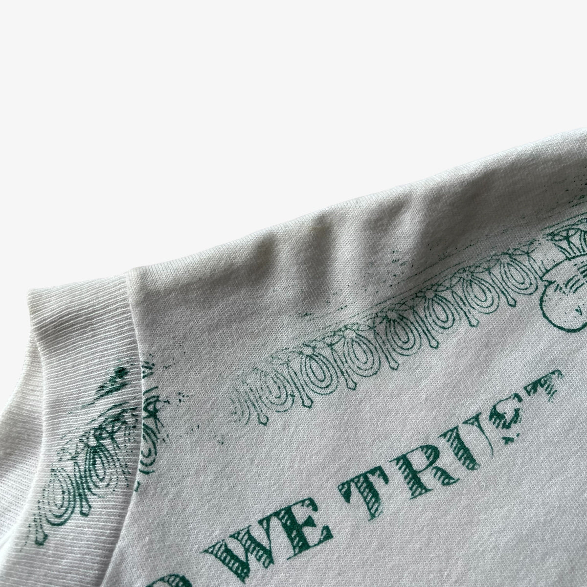 Close-up of a vintage white t-shirt, featuring a dark green distressed graphic print with the words 'WE TRUST' and decorative scrollwork.