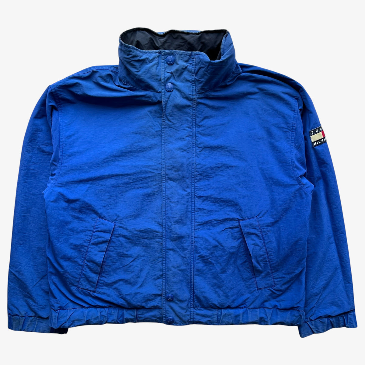 A blue sailing jacket with a high stand-up collar, front snap closure, and two front flap pockets, laid flat against a light grey background. A Tommy Hilfiger flag logo patch is visible on the left sleeve.