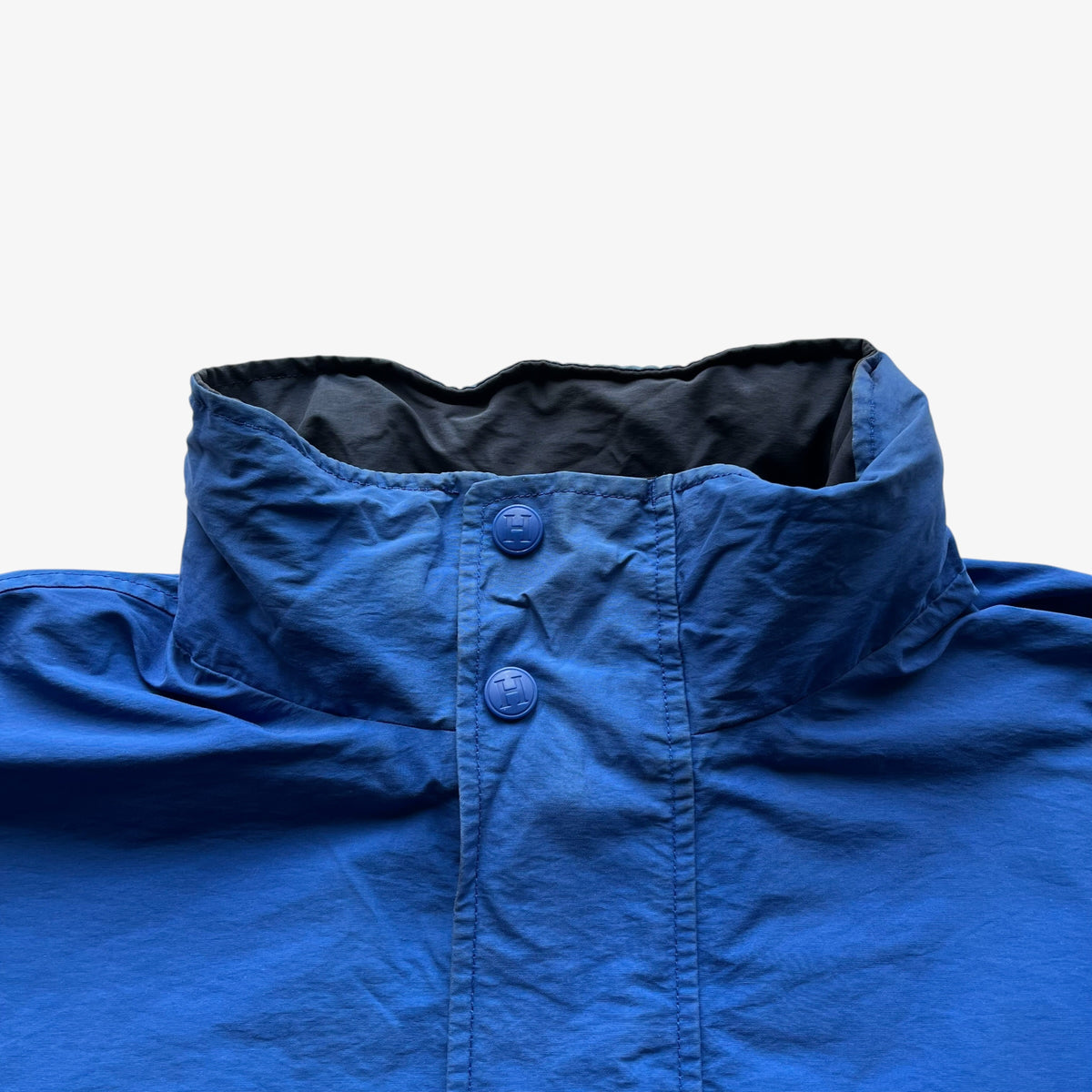 Close-up of a blue lightweight jacket with a high collar and matching blue buttons, set against a plain light grey background.