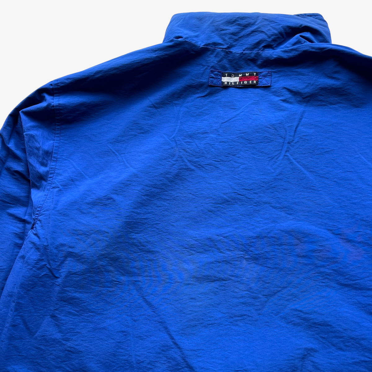 A close-up of a bright blue long-sleeve shirt laid flat, showing the back collar and a small Tommy Hilfiger flag label stitched near the neckline.