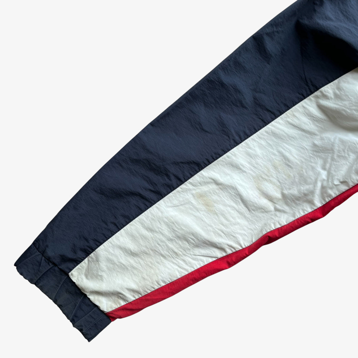 A close-up of a sleeve or fabric panel with a diagonal layout, featuring a dark navy blue upper section, a wide off-white middle section, and a thin red stripe along the lower edge.