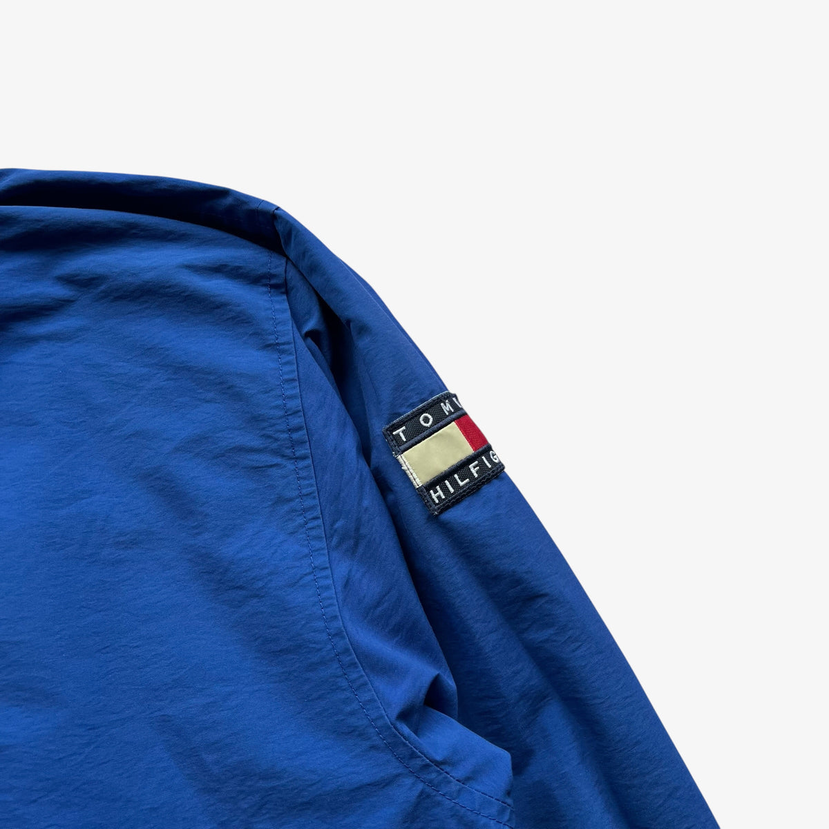 Close-up of a blue Tommy Hilfiger jacket sleeve showing the brand's embroidered flag patch on the shoulder, set against a plain white background.