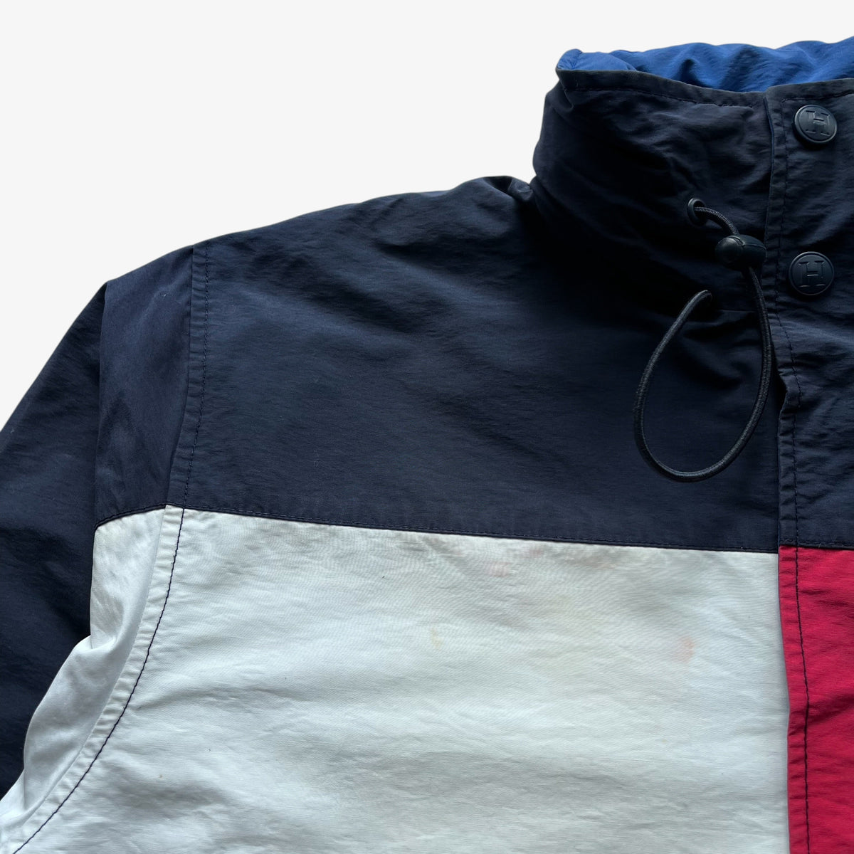 A close-up of a colour blocked windbreaker jacket with a navy blue upper section, a large off-white central panel, and a red vertical stripe on the right side, featuring a high collar and black drawstring.