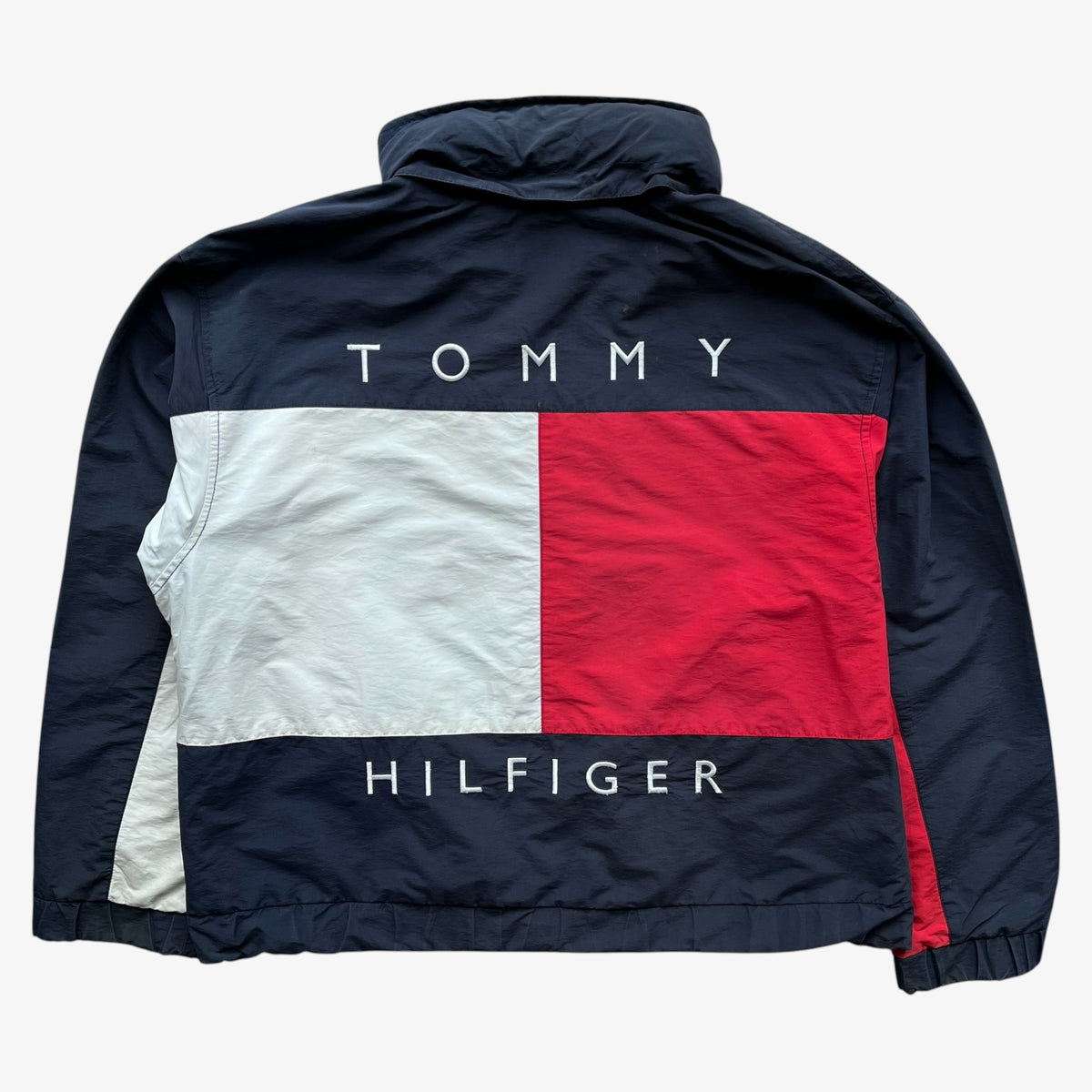 A navy blue Tommy Hilfiger windbreaker jacket shown from the back, featuring the classic red, white, and navy flag design across the back with 'TOMMY' above and 'HILFIGER' below in white text.