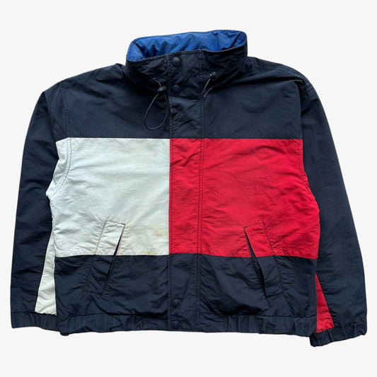 A vintage 90s Tommy Hilfiger colour block windbreaker jacket featuring a navy blue base with a large white block on the left chest and a large red block on the right chest, resembling the classic flag design. The jacket has a high collar with a blue lining, front zipper, and front pockets.