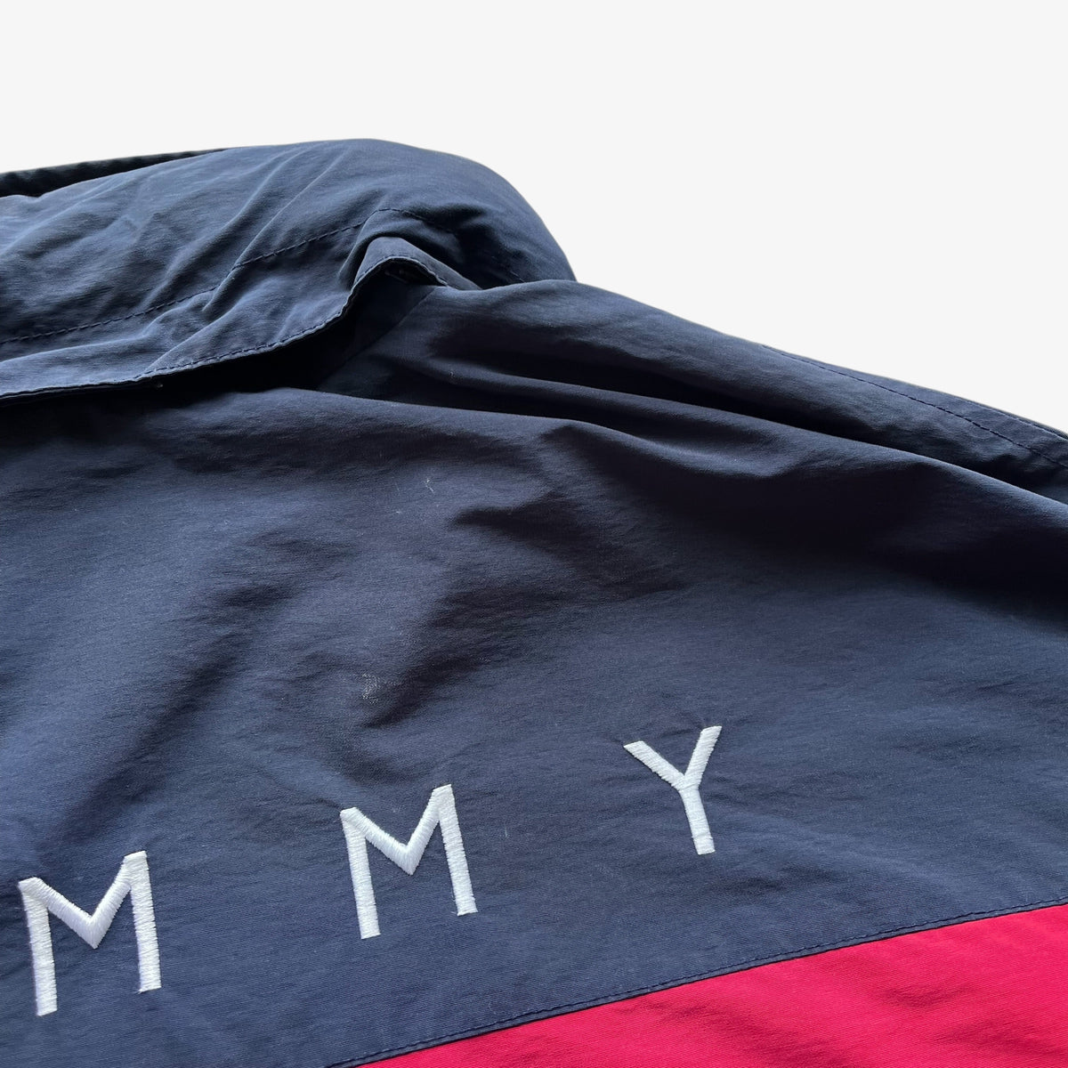 Close-up of a navy sailing jacket with white embroidered letters spelling 'Tommy Hilfiger' and a red colour block stripe at the bottom, photographed against a white background.