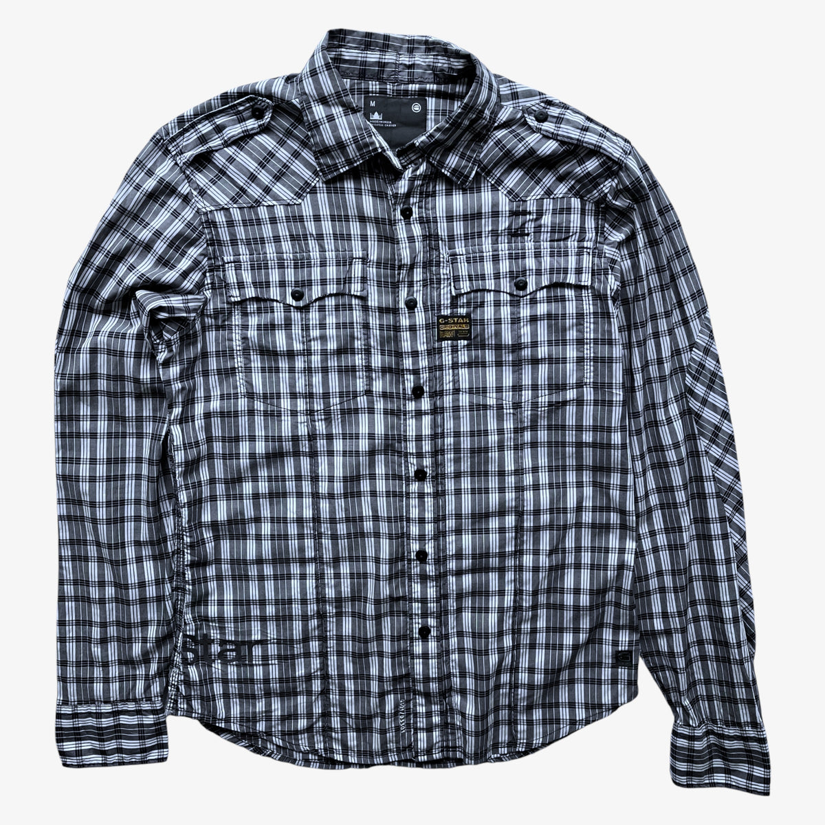 A long-sleeve G-Star plaid button-up shirt in black and white, featuring two chest pockets with button flaps and a small rectangular brand patch on the left chest, laid flat against a light grey background.