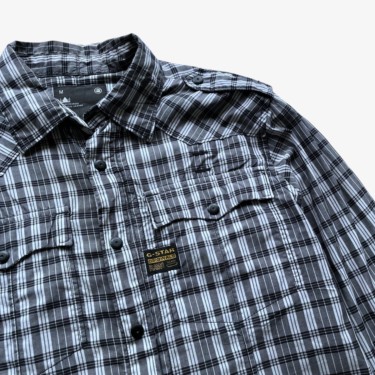 A close-up of a G-Star men's plaid button-down shirt in black, white, and grey, featuring a chest pocket with a flap and a visible G-Star brand patch.
