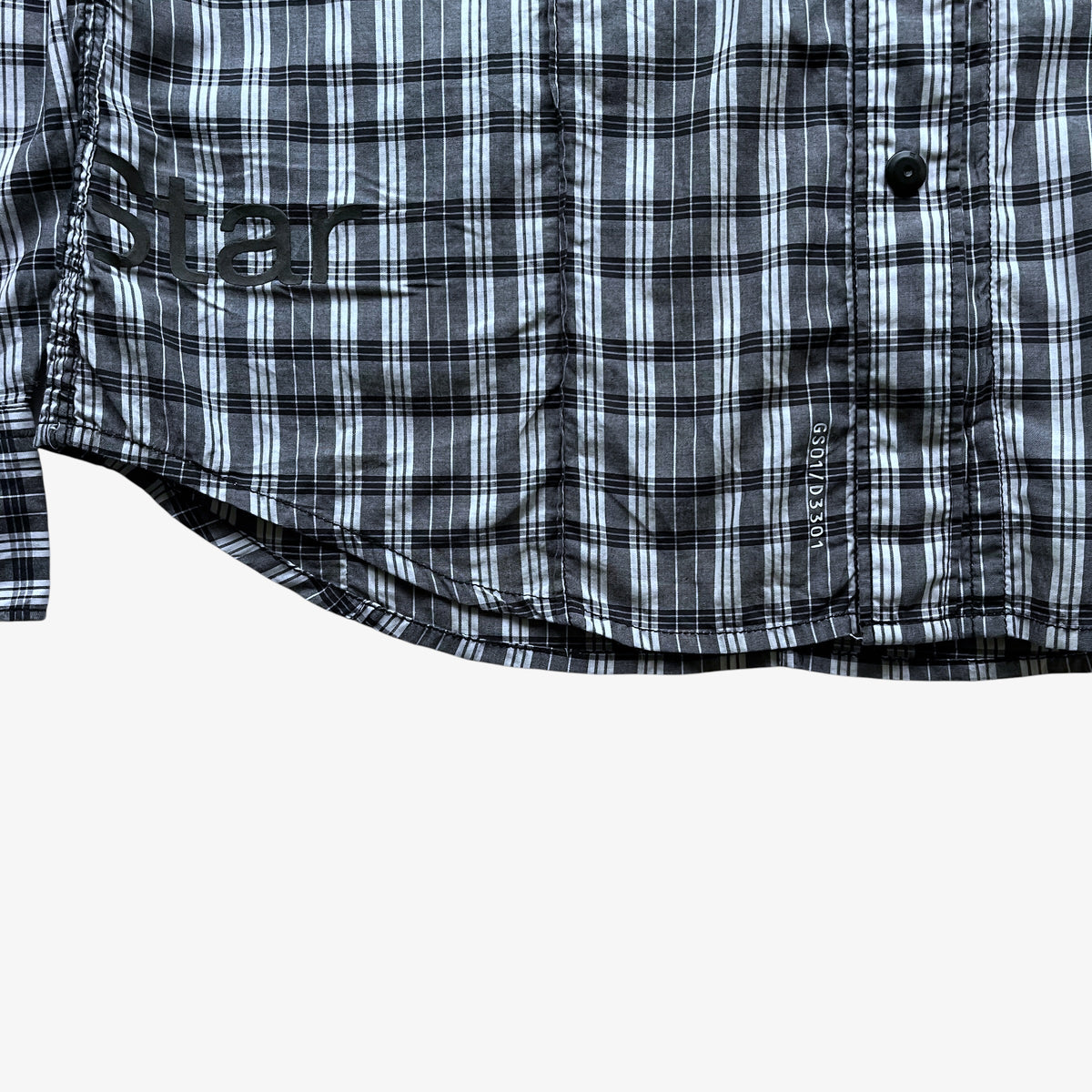 Close-up of a plaid button-up shirt in black, white, and grey, showing the fabric pattern, a button, and a small vertical brand tag on a white background.