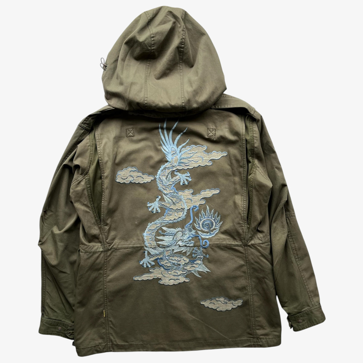 A khaki green Maharishi hooded jacket shown from the back, featuring a large light blue embroidered dragon design with clouds across the centre back.